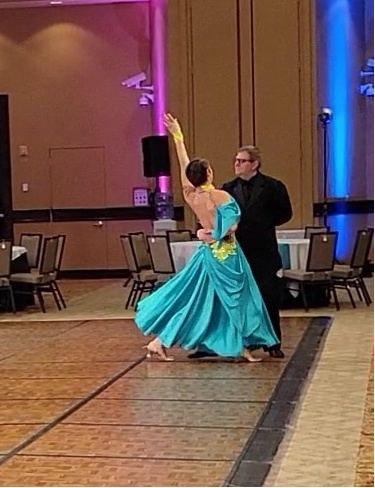 Marlena in a teal ad yellow dress doing a corte in American Tango with a partner in a black suit