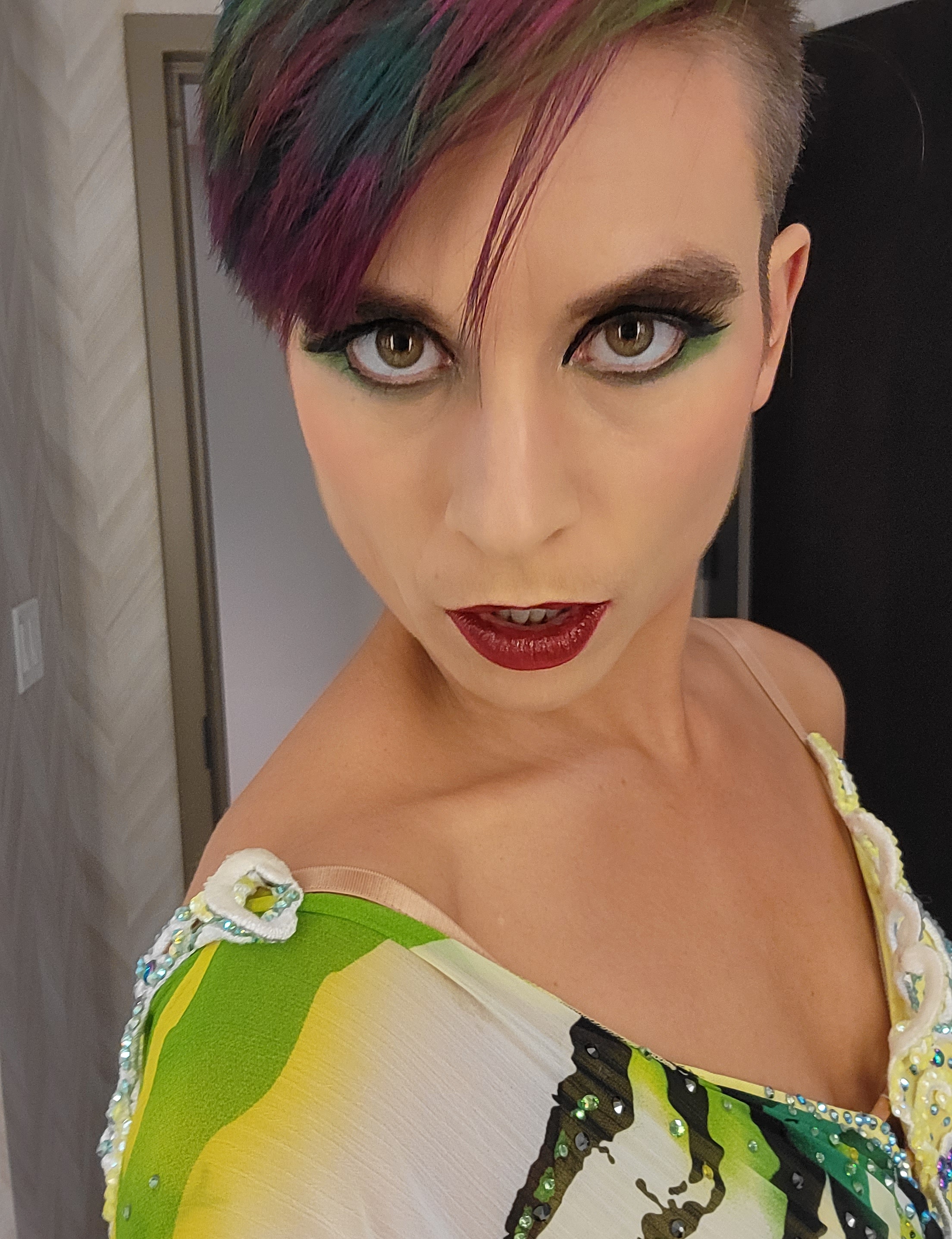 Marlena, a nonbinary person with deep pink, purple, and green hair and green eyeshadow, wearing a green and black striped dress, staring fiercely into the camera