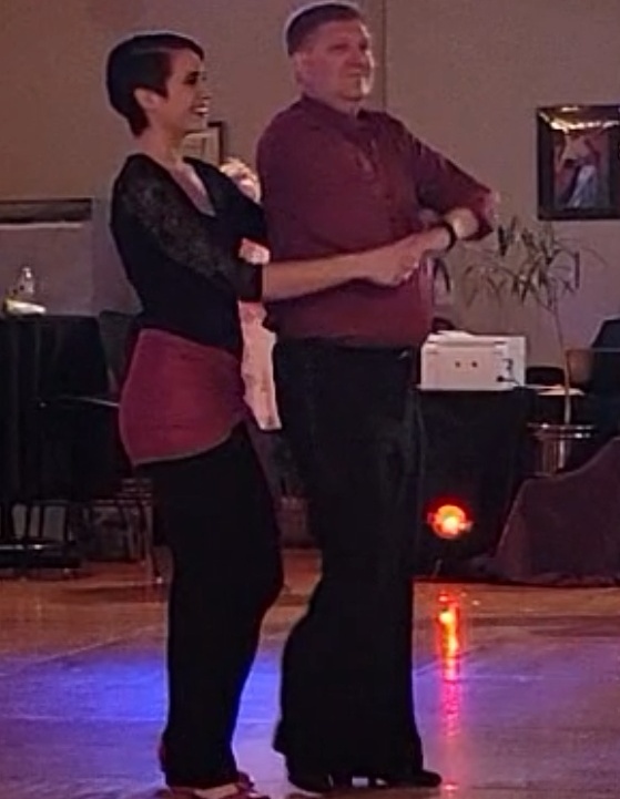 Marlena, a nonbinary person in a black outfit and pink hip wrap, leads a man in a pink shirt and black pants