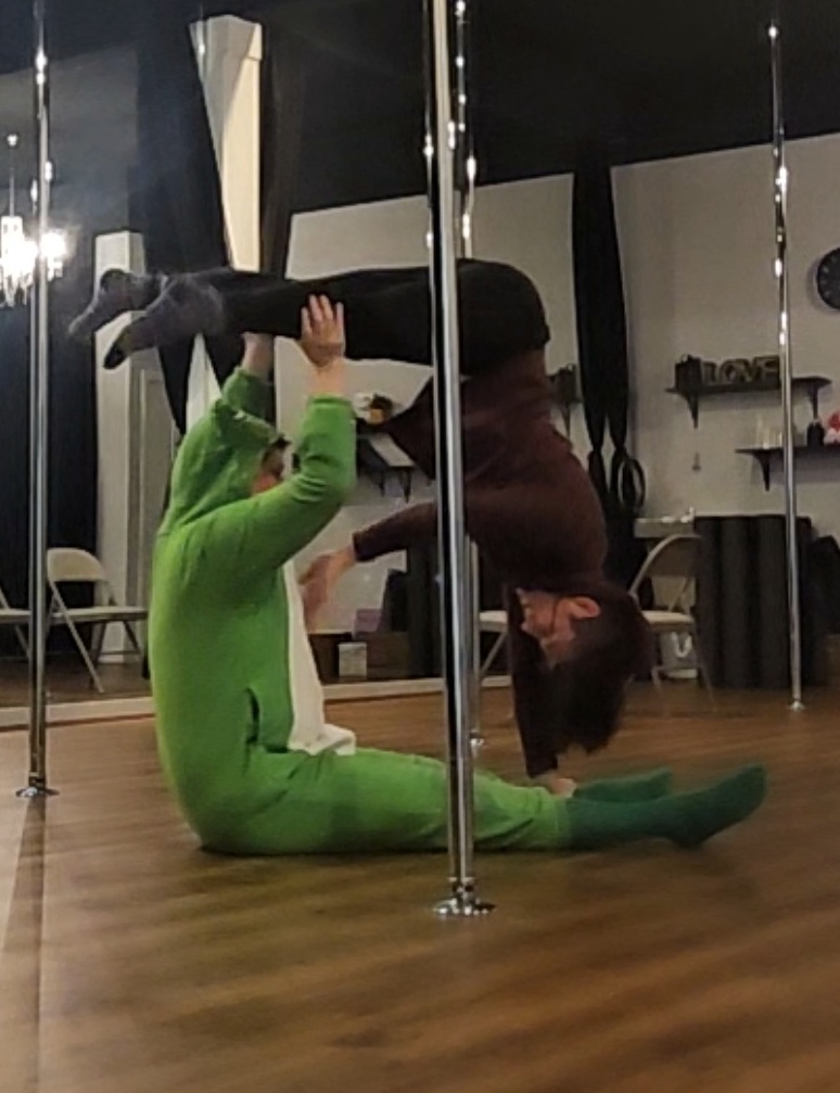 Marlena, dressed in red and black as a scorpion, is upside-down in the square yoga pose, supported by dance partner dressed in a frog onesie.