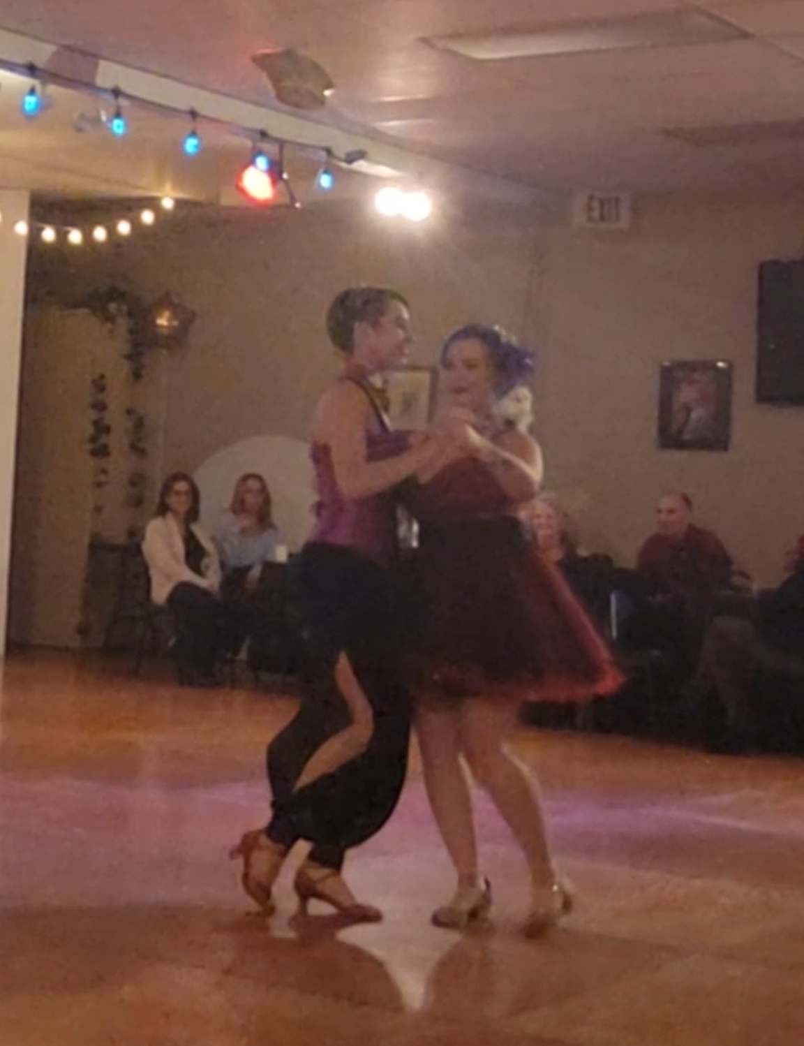 Marlena, a nonbinary person in a corset and pants, dances with a woman in a dress. They both wear bright costumes