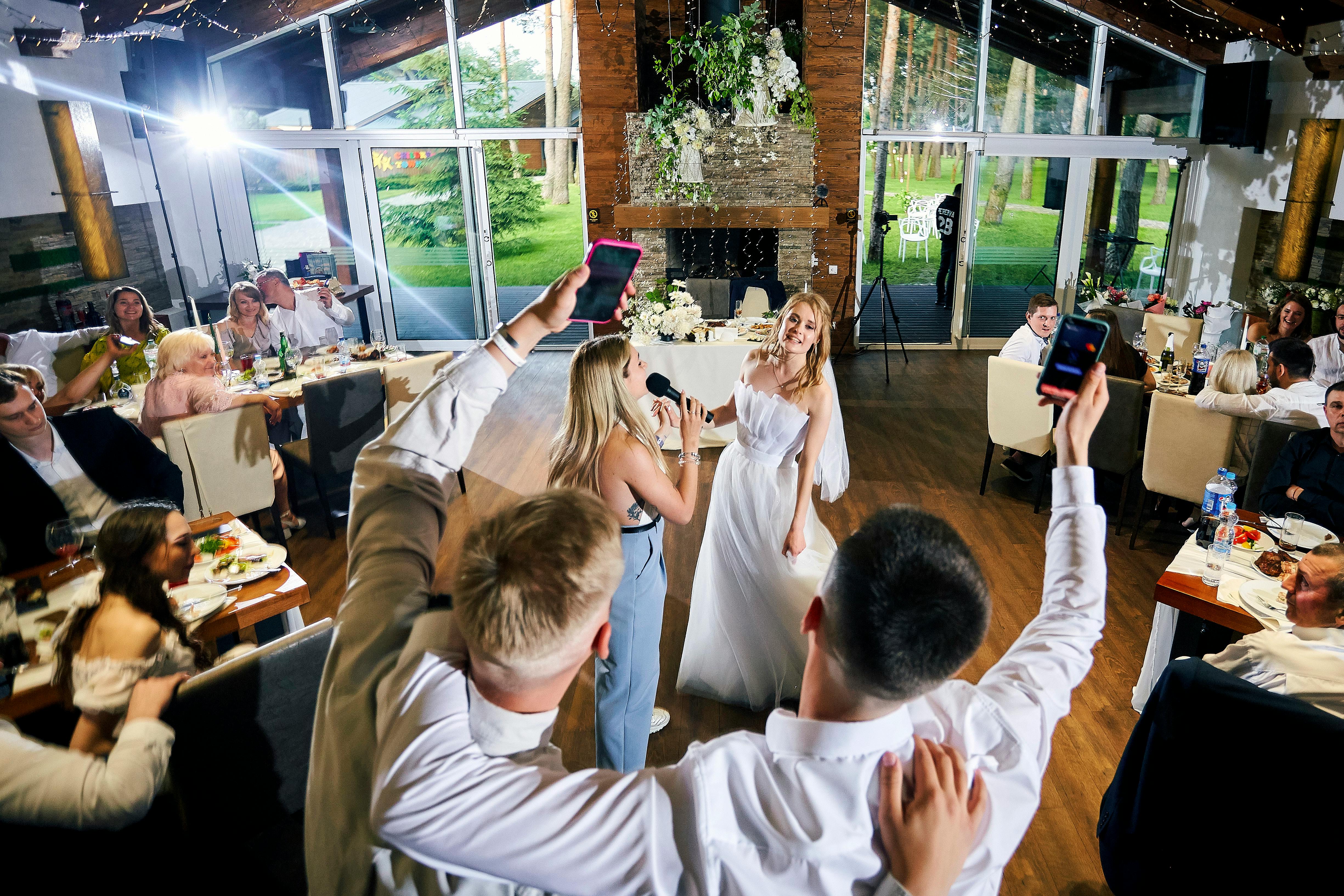 Two white women, one in a white wedding dress, and one in a blue dress, stand on the dance floor amidst seated wedding guests.