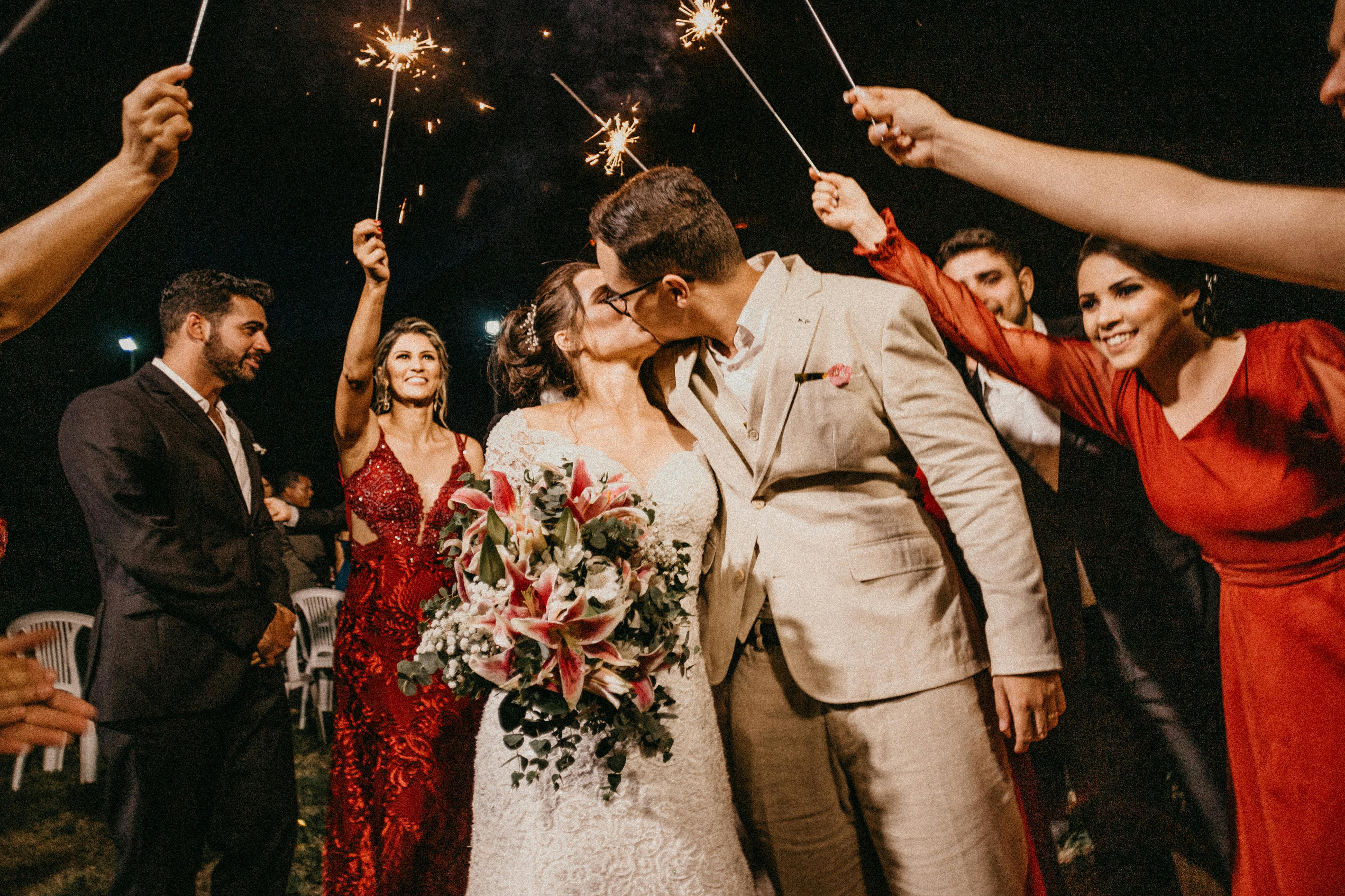 A woman in white dress holding flowers and person in tan suit kiss, surrounded by friends holding sparklers.