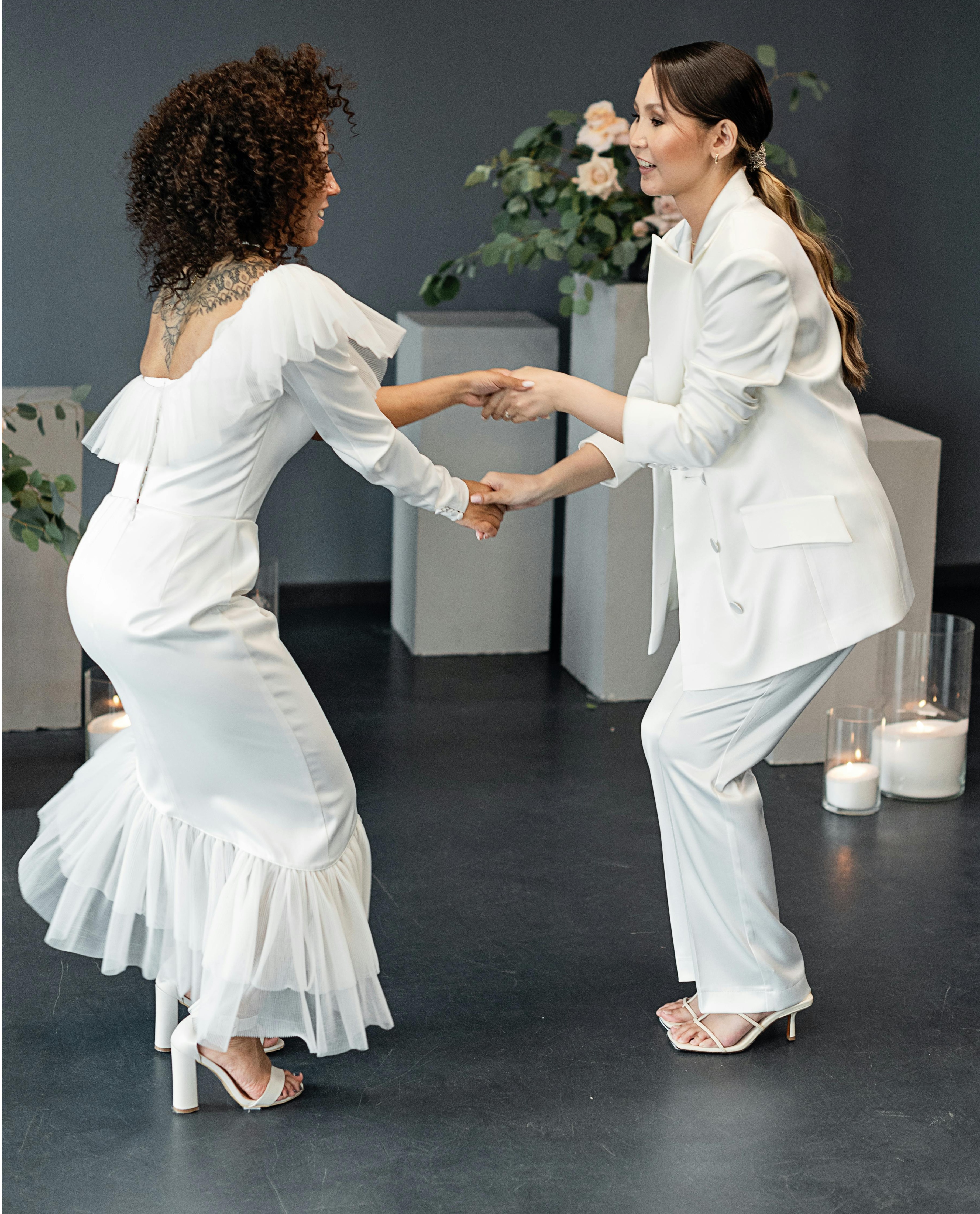 Two BIPOC women, one in a white dress, the other in a white suit, dancing together in front of flowers on tables.