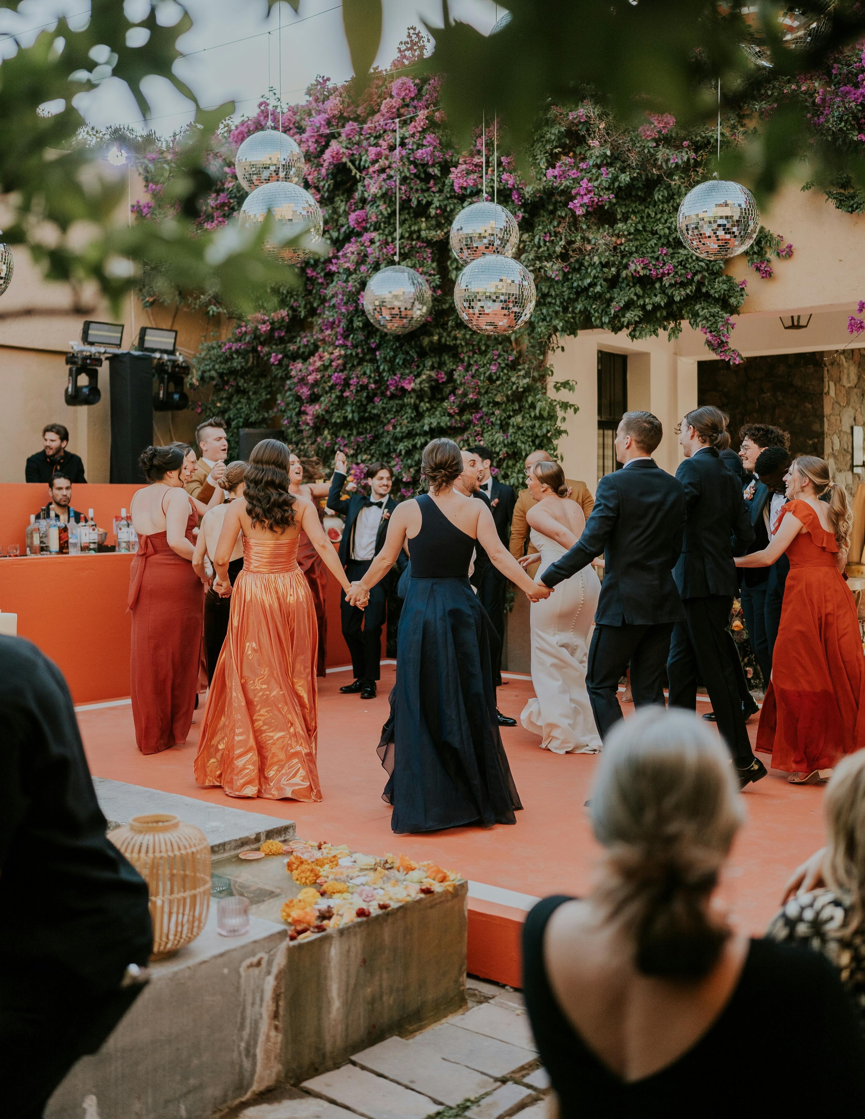 About ten people of various genders, wearing suits and floor-length dresses, dance hand-in-hand around a wedding couple.