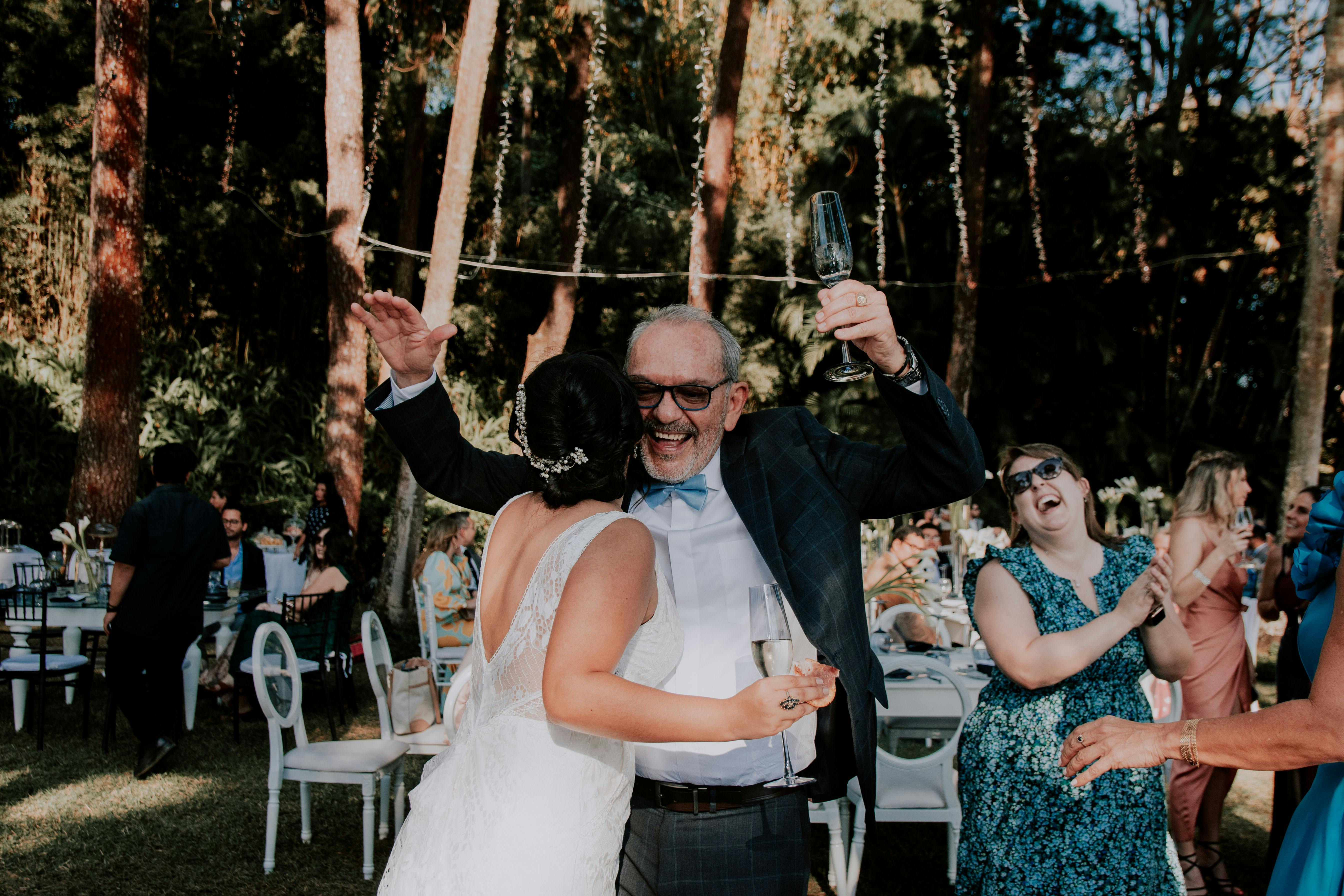 Wedding guests in dresses and suits stand around clapping and dancing. At the front, a woman in a wedding dress whispers into the ear of a man. They are dancing and holding cocktails.